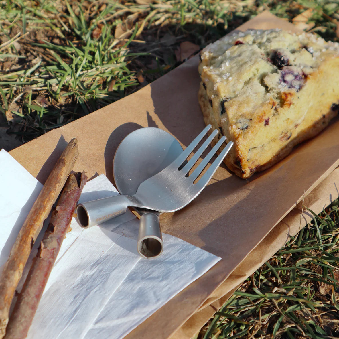 Huckleberry Forest Cutlery stainless steel fork, spoon, and sticks displayed with a slice of berry bread outdoors on paper.