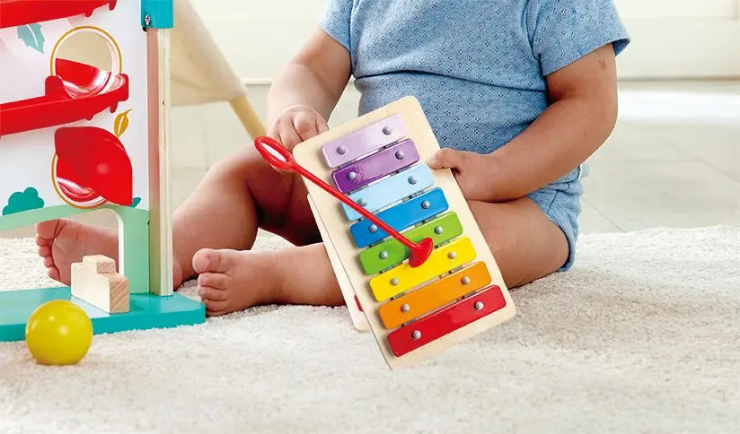 Child playing with the Melody Mansion Ball Run holding the rainbow xylophone on a soft carpet.