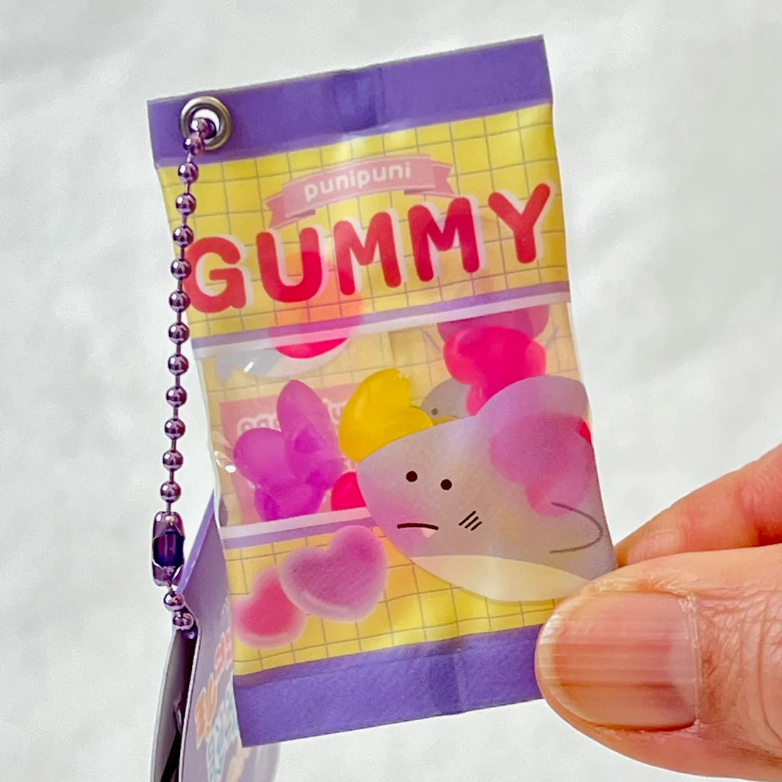 Hand holding a colorful Gummy Shark Keychain resembling a small bag of squeezable gummy toys with a purple chain.
