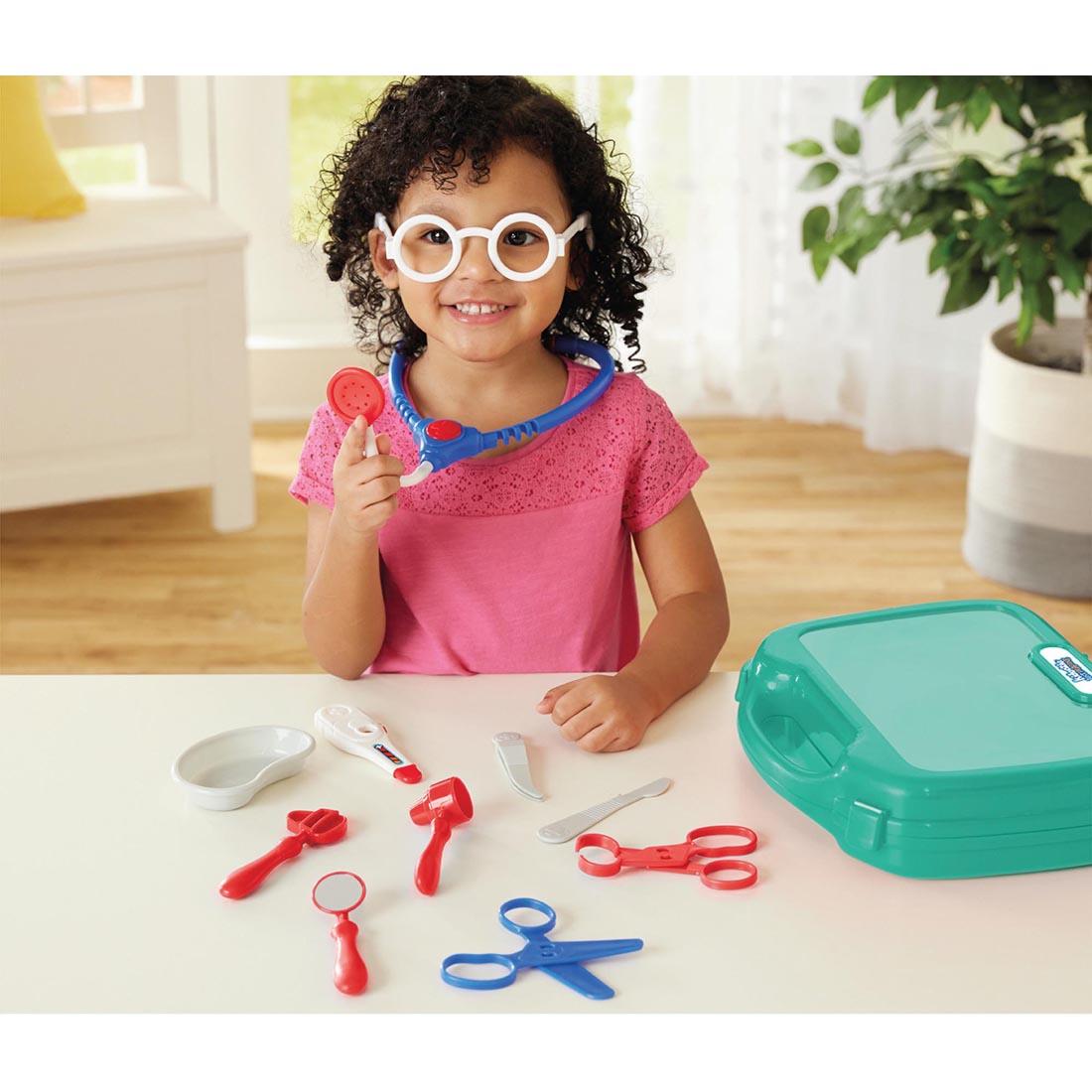 Smiling child playing with Doctor Kit toy set including stethoscope, thermometer, scissors, and medical case on table.