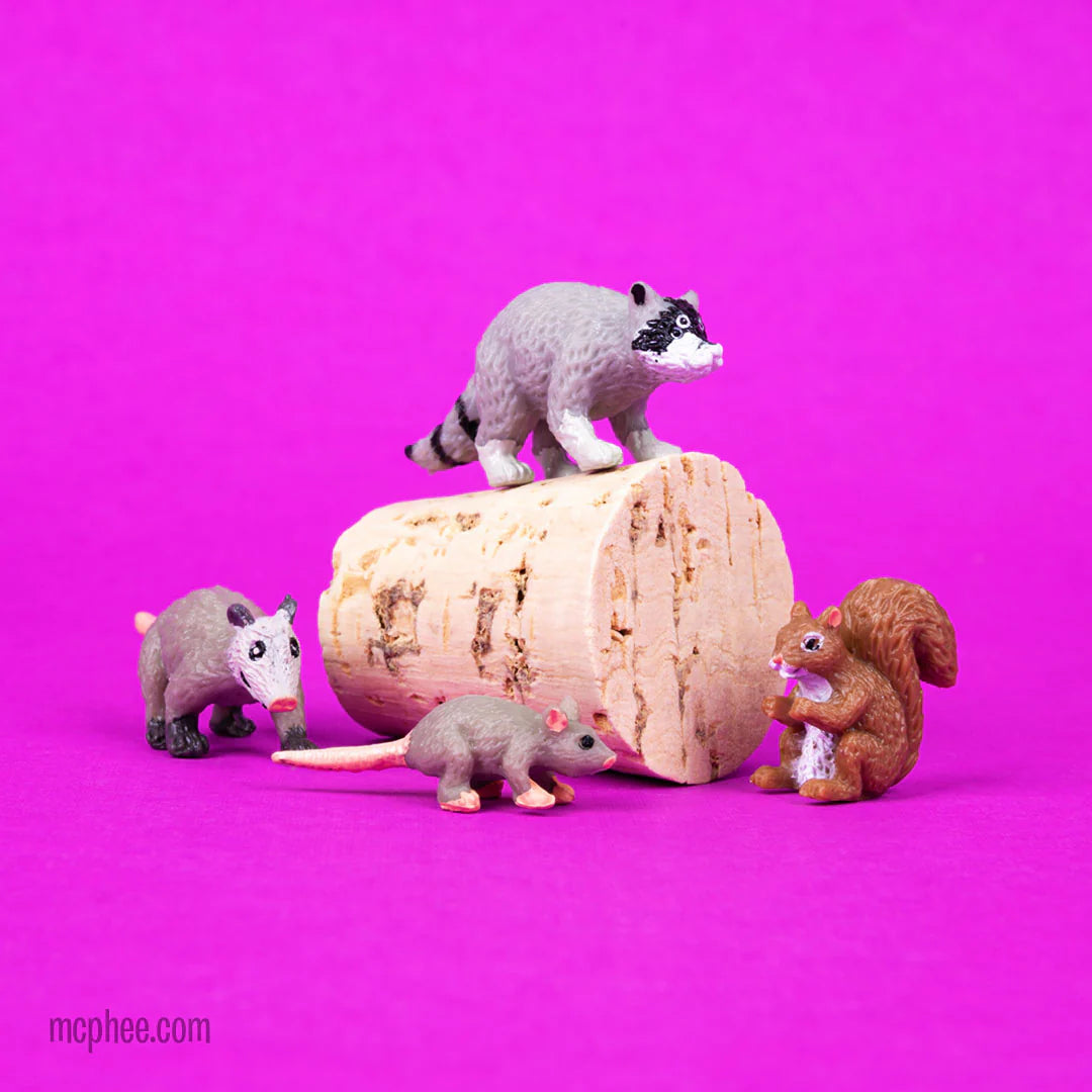 Small detailed vinyl models of city animals including raccoon, squirrel, rat, and opossum on a wooden log against pink background