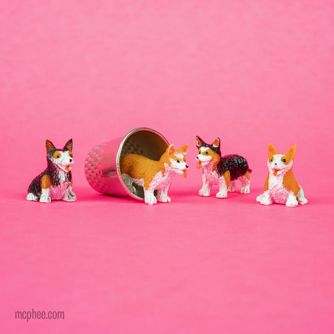Four small soft vinyl Itty Bitty Corgis in two poses and color combos with a metal cup on a pink background.