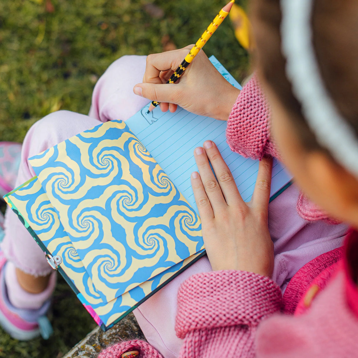 Person writing in the open Axolotl Diary journal with a blue and yellow swirl patterned inside cover outdoors.