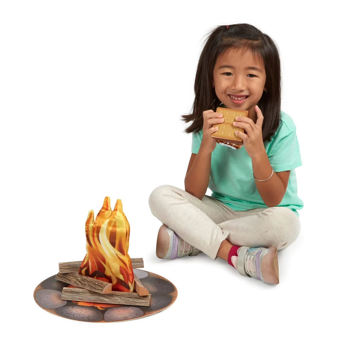 Girl sitting and smiling while holding a s'more next to the Let's Explore Campfire S'mores Play Set with flame and logs.