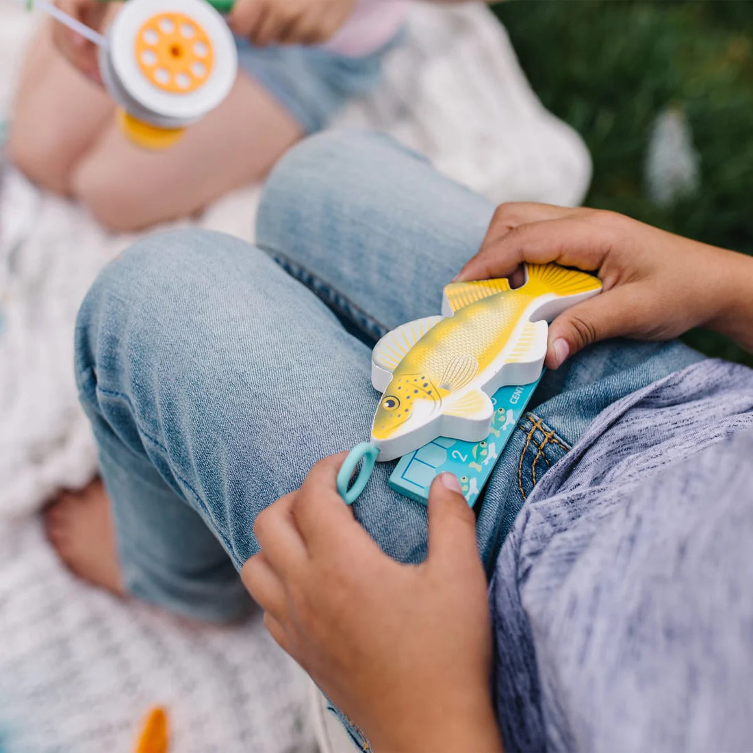 Child playing with wooden fish and fishing rod from the Let's Explore Fishing Play Set on a blanket outdoors.