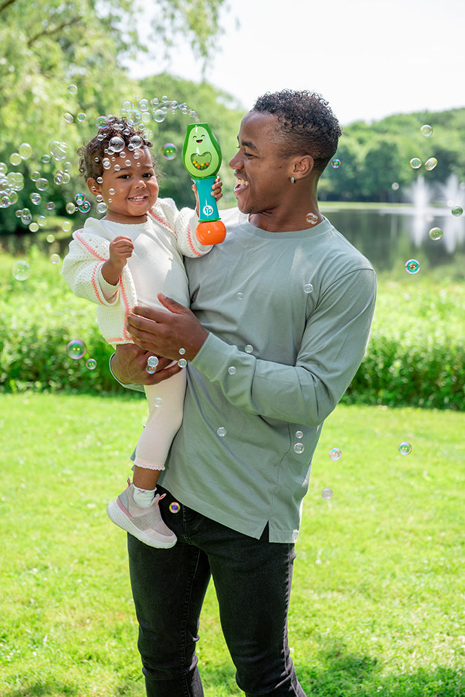 Man holding toddler playing outdoors with Fisher Price No-Spill Shake & Go Bubble Maraca blowing bubbles.