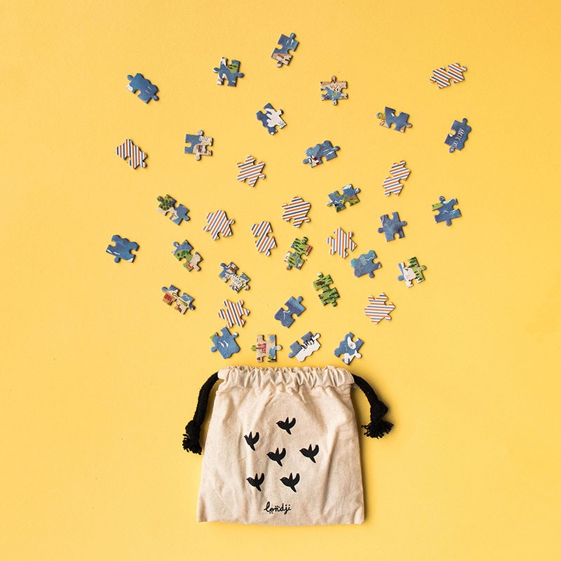 100pc Discover the World Pocket Puzzle pieces scattered above a small drawstring fabric pouch on yellow background