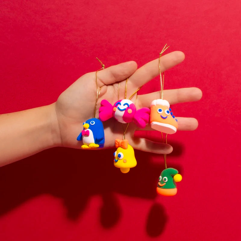 Hand holding colorful DIY Magic-Do Clay Ornaments including penguin, candy, boot, bell, and elf shapes on gold strings.