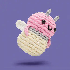 Small crocheted firefly toy with a pink head and cream body, featuring black eyes and white wings on a purple background