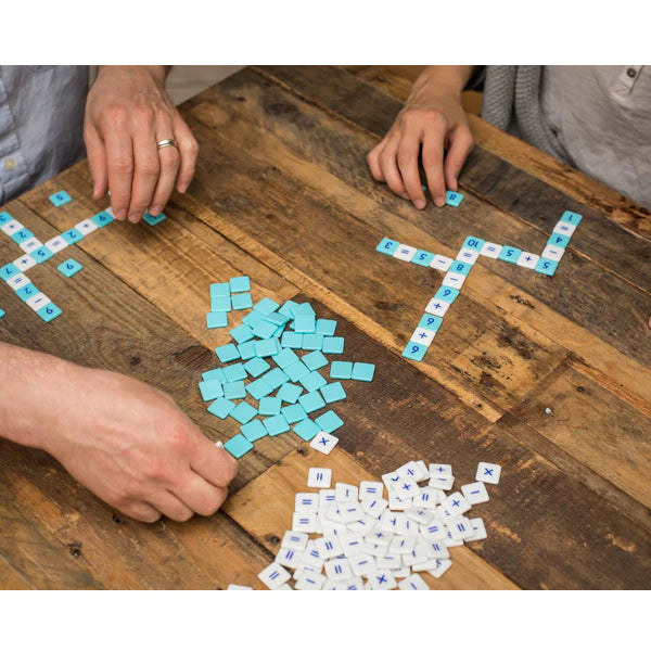 Hands playing Mobi Number Tile Game on a wooden table with blue and white math equation tiles arranged crossword-style.
