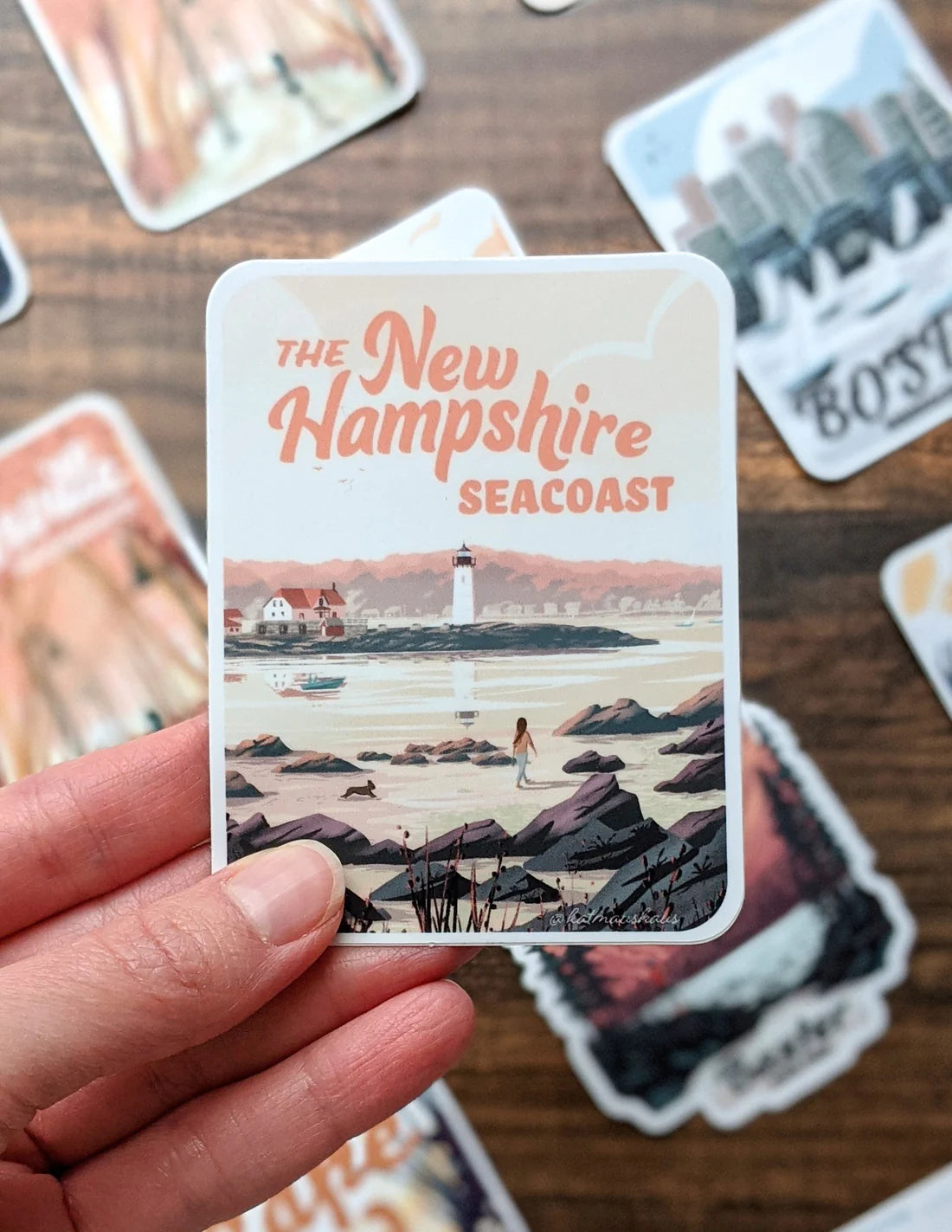 Hand holding New Hampshire Seacoast Sticker featuring lighthouse and rocky shore illustration with soft colors.