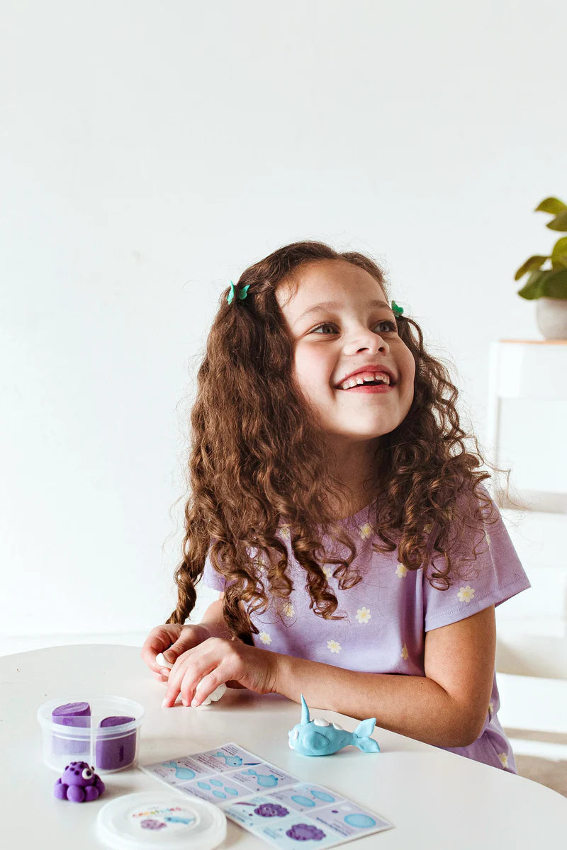 Girl smiling and playing with clay characters made from Creatibles Air Dry Clay Kit Ocean BFFs on a white table.
