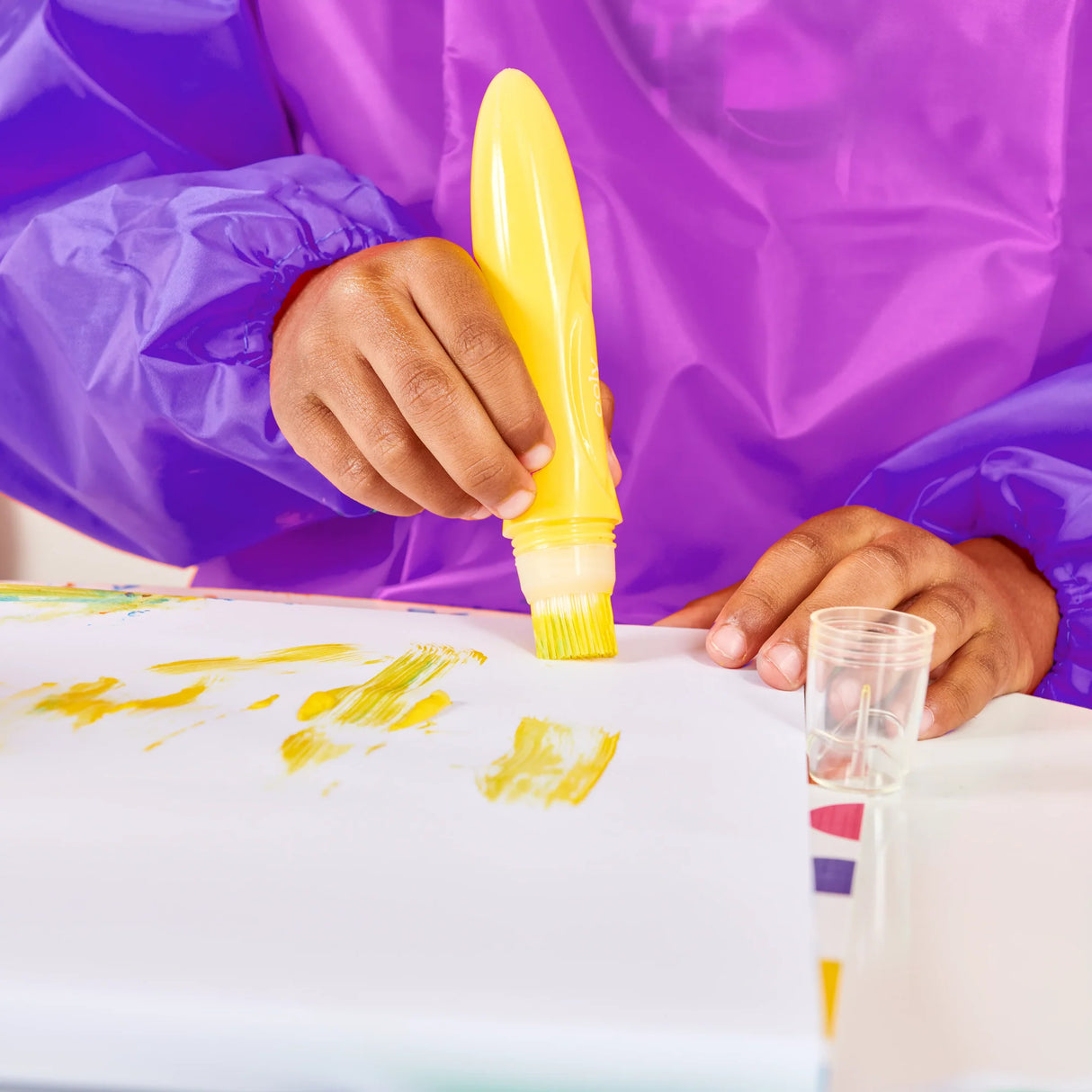 Child using a yellow Easy Squeezy Squeezable Poster Paint Brush to apply paint on paper, wearing a purple smock.