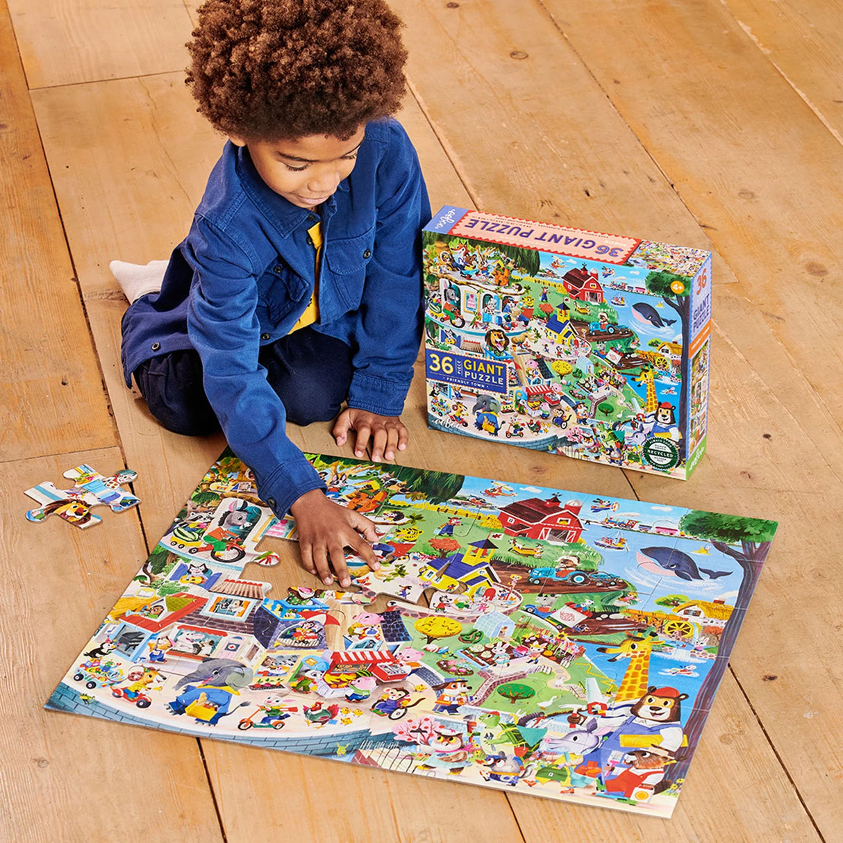 Child assembling 36pc Friendly Town Giant Puzzle showing colorful animals and community scene on wooden floor.
