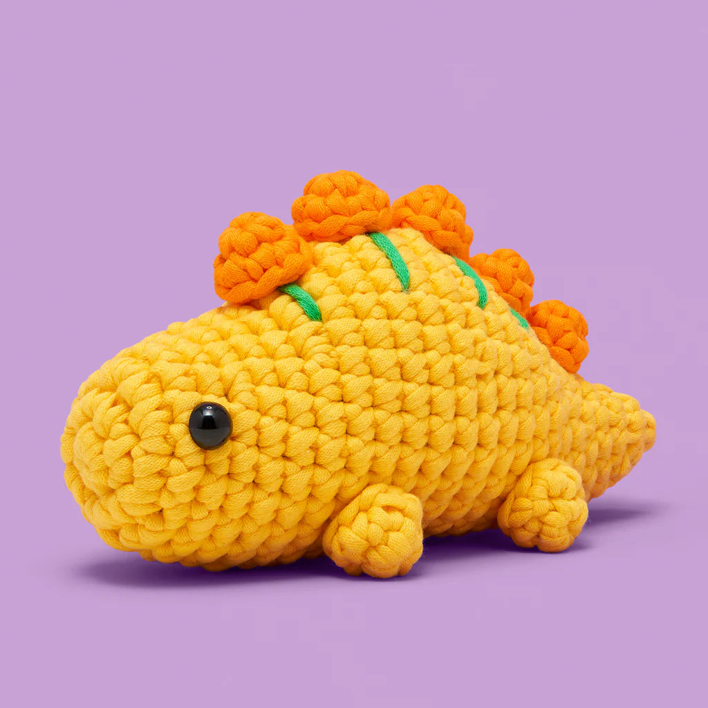 Cute crocheted yellow Stegosaurus with orange plates and black eyes on a purple background