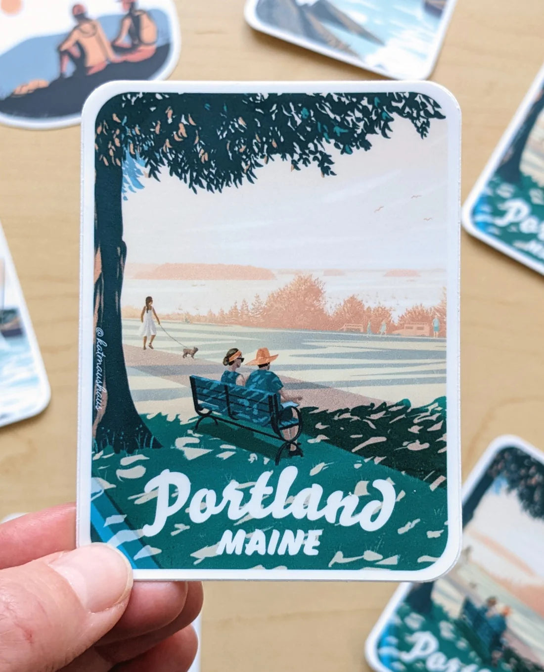 Hand holding Portland Maine Bench Sticker showing a scenic park illustration with people on a bench under a tree.