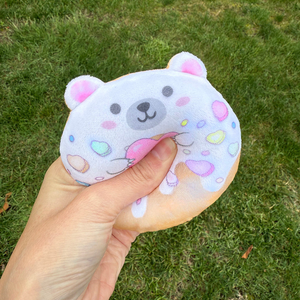 Hand squeezing a soft plush toy shaped like a donut with a cute bear face and colorful sprinkles outdoors on grass.