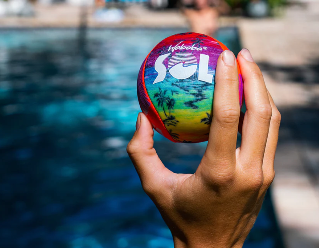 Hand holding colorful Waboba SOL Ball with palm tree design next to a swimming pool on a sunny day.
