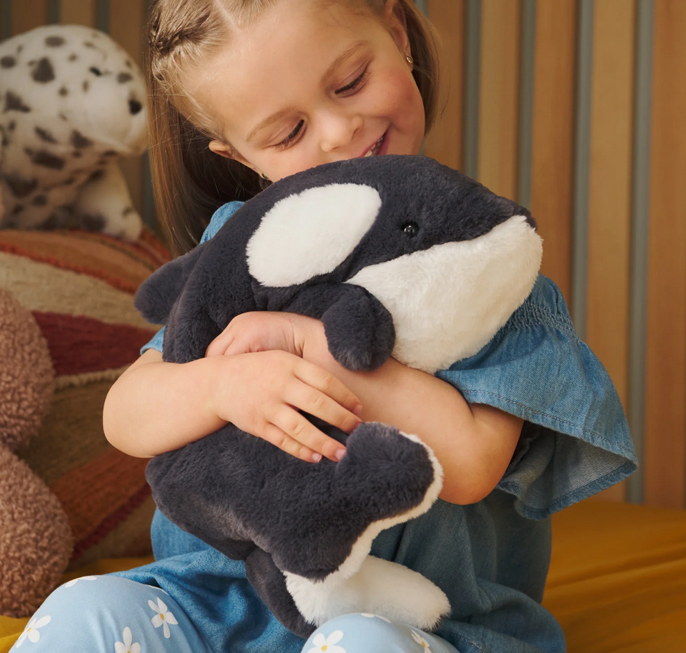 Child smiling and hugging a soft, black and white plush toy Snuffles Orca Flynn sitting upright.