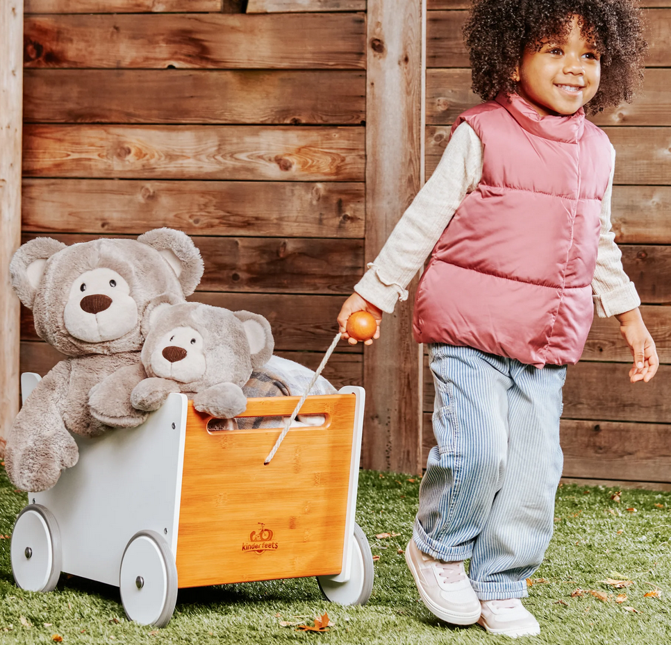 Teddy Bear Mukki plush toy with woodgrain-inspired fur sitting in a wooden wagon pulled by a smiling child.