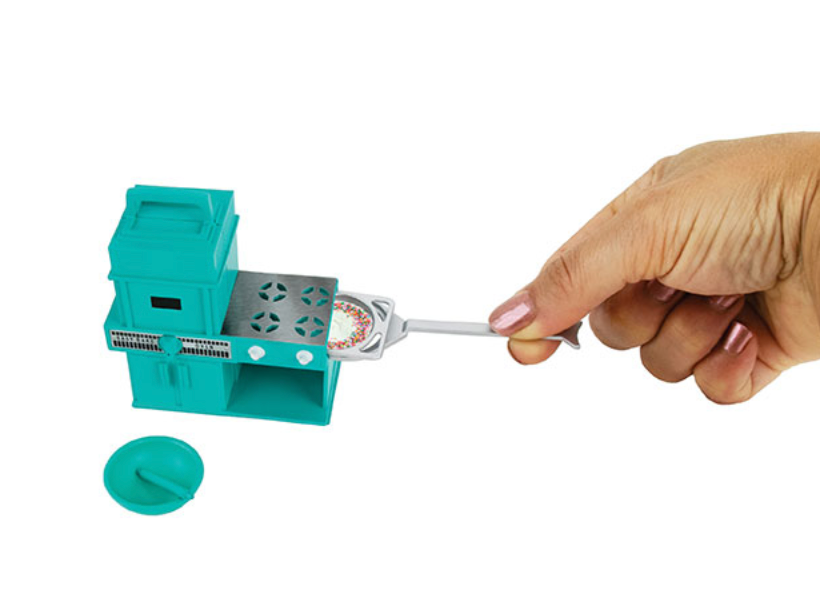 Hand holding mini spatula with tiny cake next to turquoise World's Smallest Easy Bake Oven toy set.