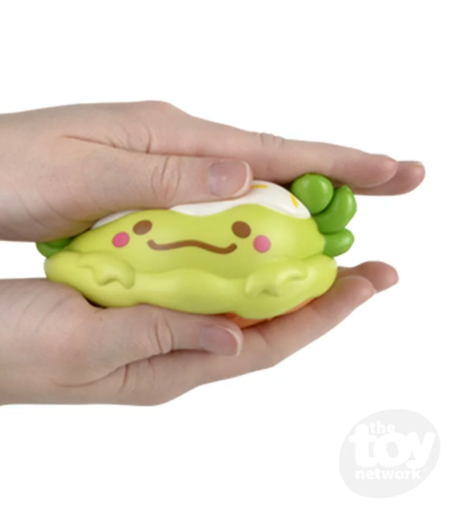 Hand holding a yellow 5-inch Squishy Axolotl Foodie Friend slow-rising foam toy with a smiling face and green accents.