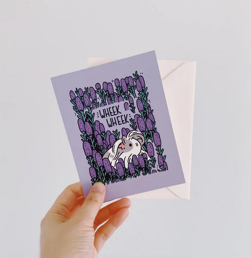 Hand holding a Guinea Pig Wheek Wheek! Card with a purple floral design and white envelope in background.