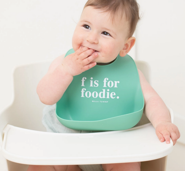 Smiling baby wearing a teal silicone bib with white text sitting in a white high chair against light background