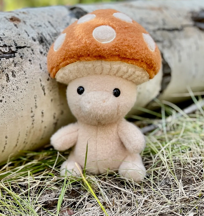 Small plush mushroom character with an orange cap and white spots sitting on grass beside a birch log