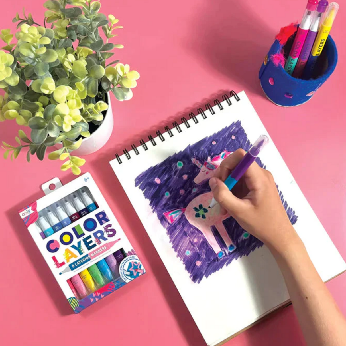 Color Layers Layering Markers shown with a hand coloring a unicorn drawing on a sketchpad on a pink surface.