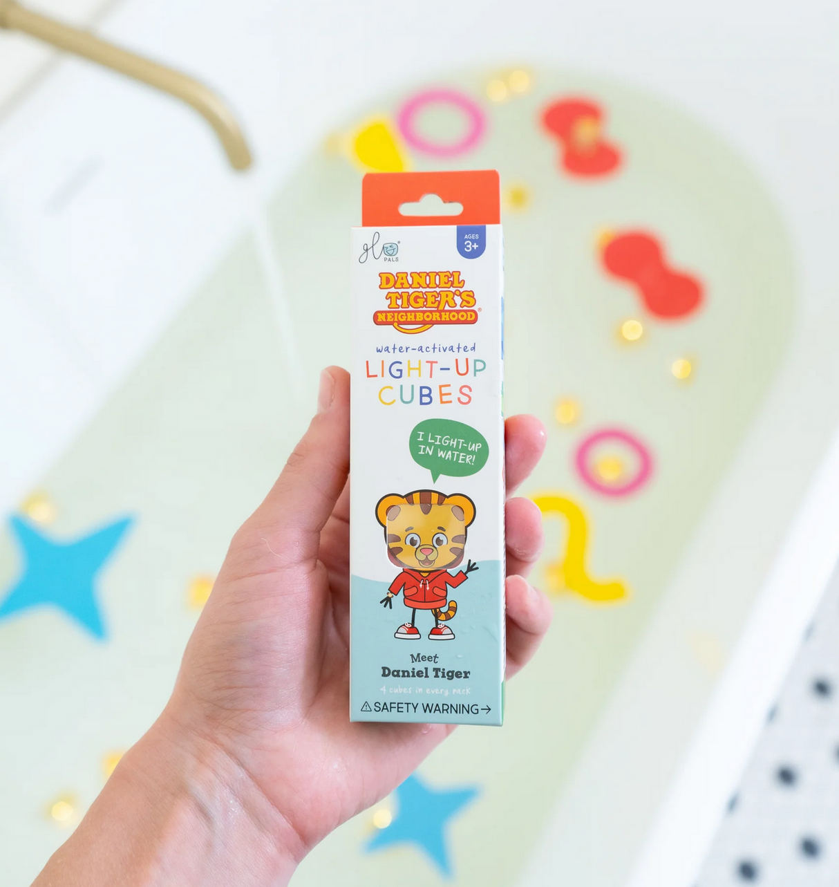 Hand holding Glo Pals Daniel Tiger's Neighborhood water-activated light-up cubes packaging over colorful bath toys background.