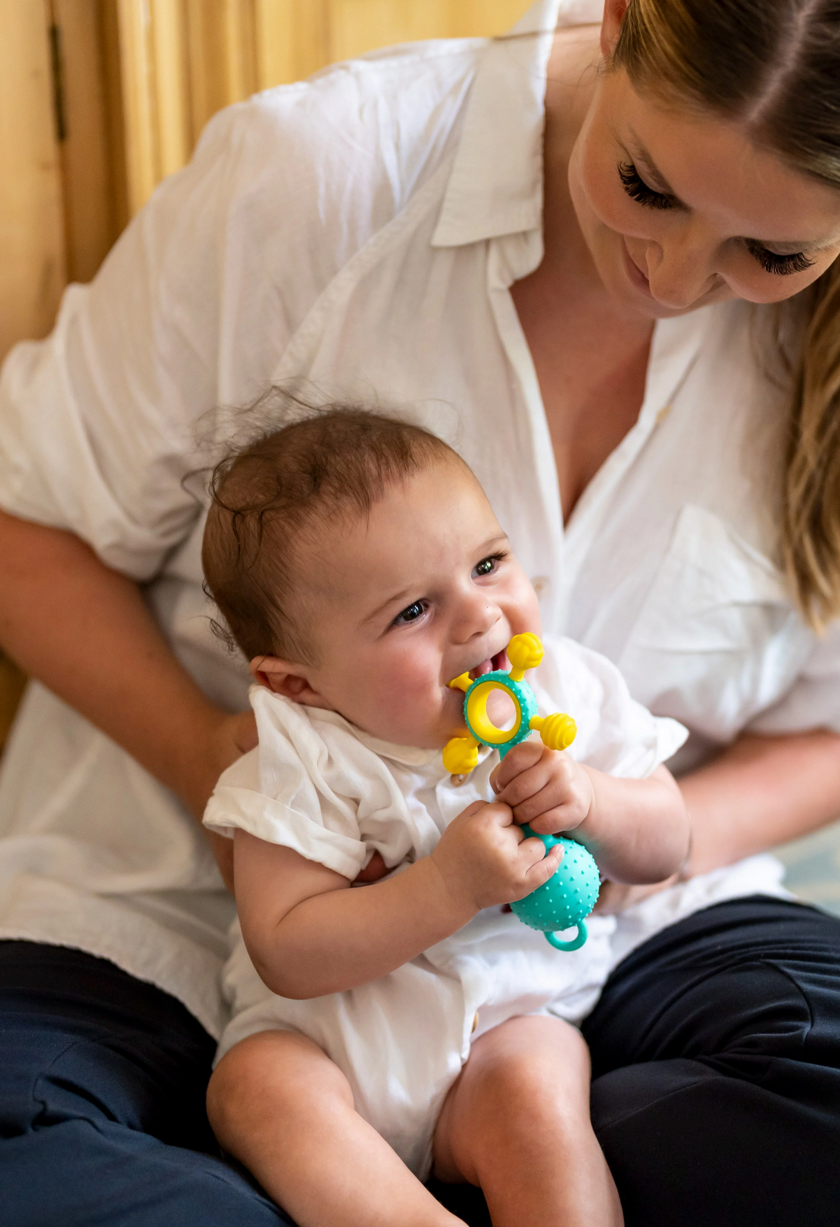 Baby chewing on Gumlii Sensory Teether + Rattle held by smiling mother wearing white shirt.