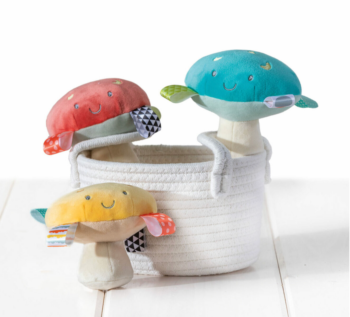 Taggies Fun Guys Mushroom Rattle in red, blue, and yellow with embroidered faces and sensory ribbons for babies.