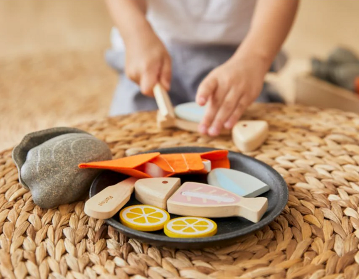 Child playing with a PlanToys Seafood Platter wooden toy set featuring fish, lemon slices, and utensils on a plate.