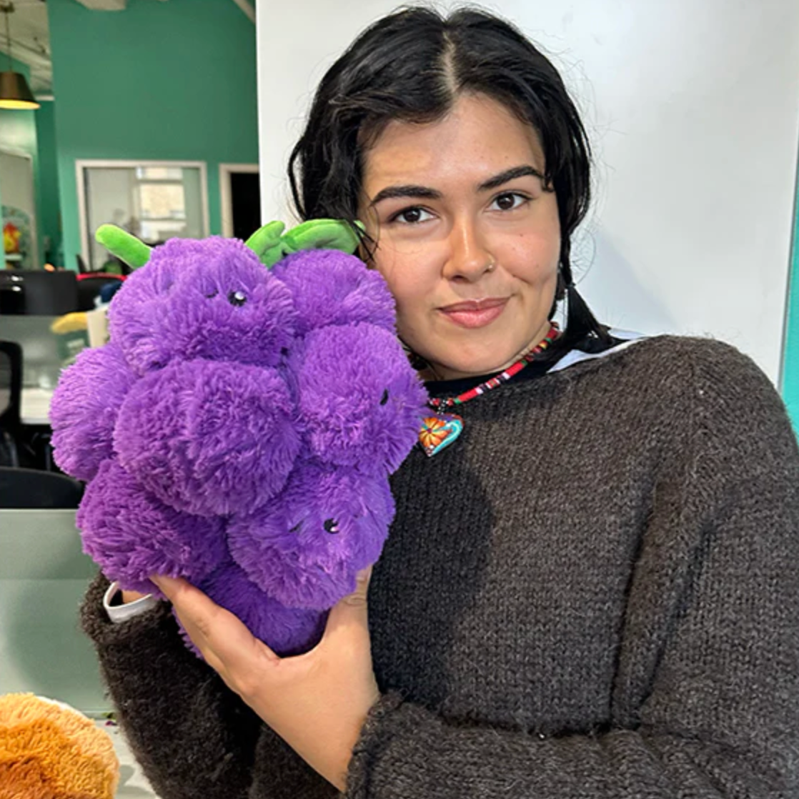 Woman holding a plush purple bunch of grapes toy called Grapes Mini in a cozy indoor setting.