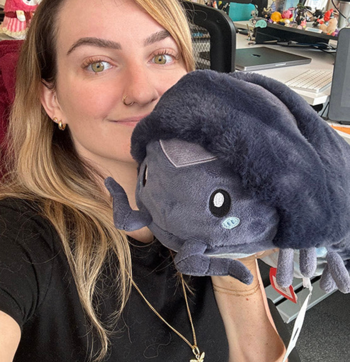 Person holding a soft plush Pillbug Mini toy with a dark shell and cute face in an office setting.