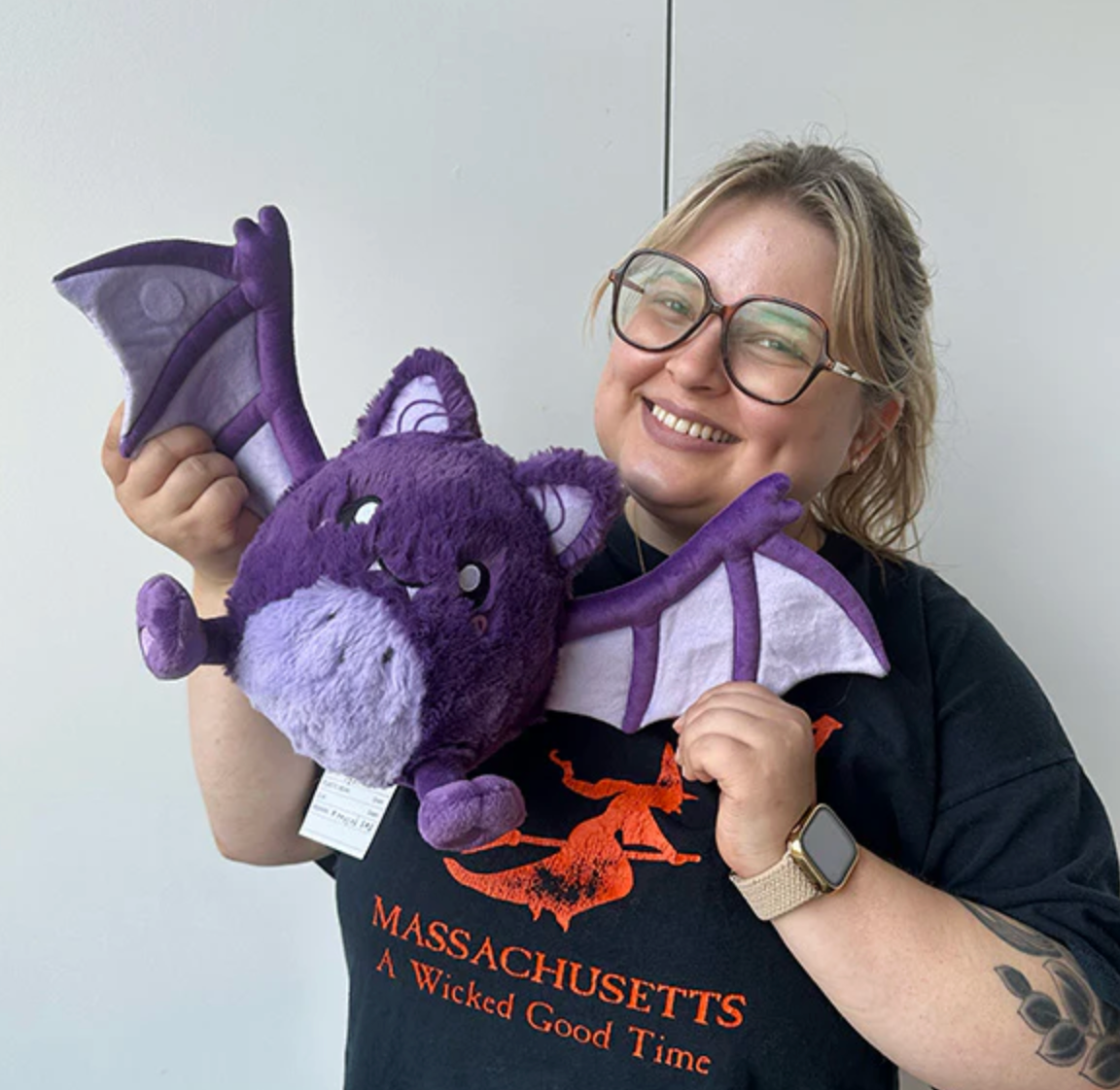 Smiling person holding a purple Spooky Bat Mini plush with outstretched wings against a plain background.