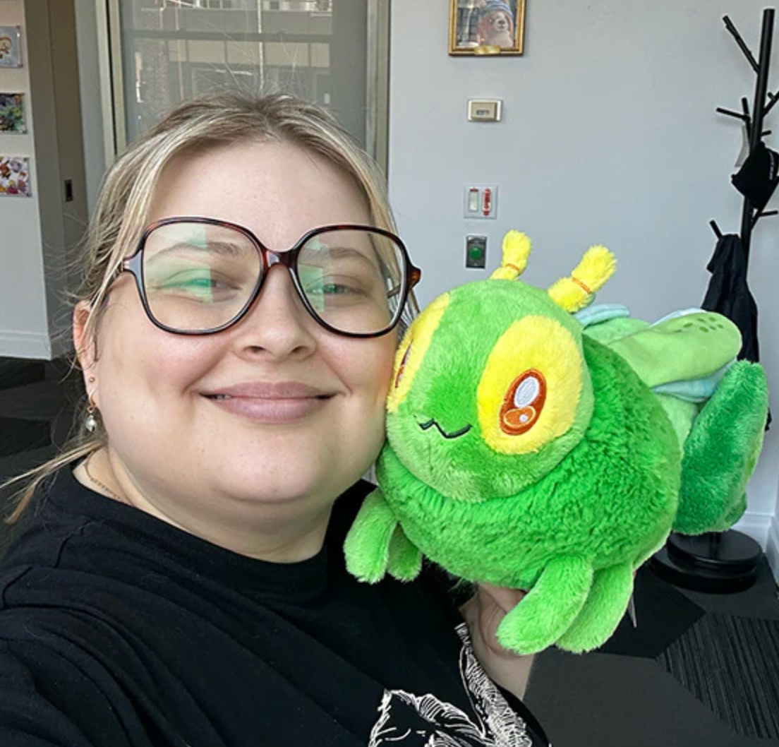Person smiling and holding a plush green Grasshopper Mini toy with yellow eyes and blue wings indoors