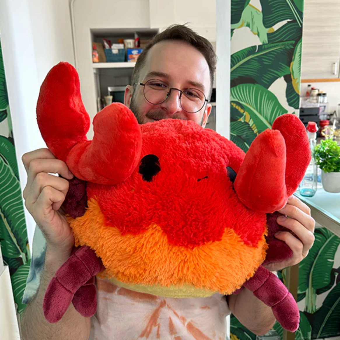 Person holding a large, colorful plush Crab with bright red claws and orange body, smiling in a cozy room.