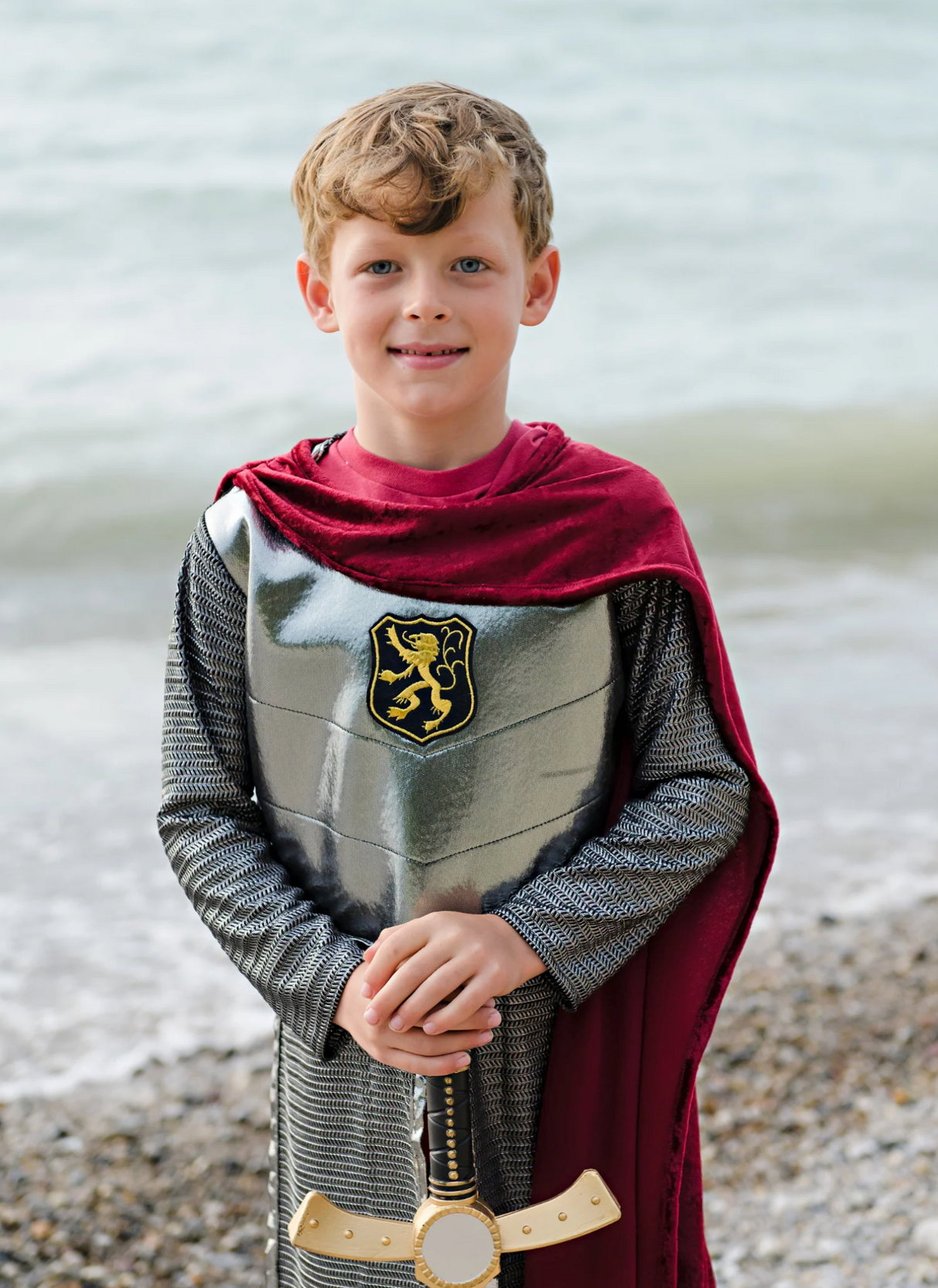 Young boy wearing the Silver Knight with Cape tunic with silver armor, red velour cape, and holding a sword by the beach.