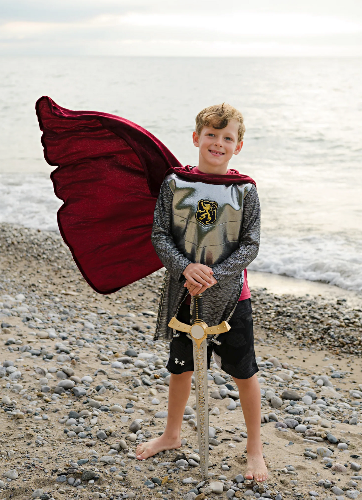 Young boy standing on rocky shore wearing Silver Knight with Cape costume holding a decorative sword.