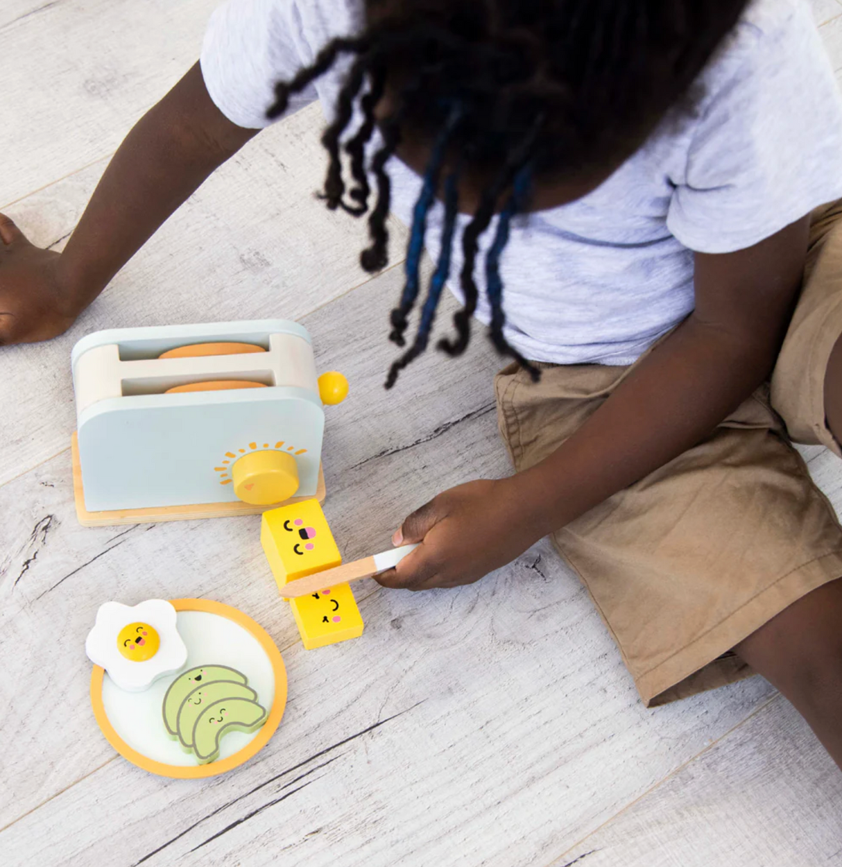 Child playing with Brunch Time Wooden Toaster Set including toaster, toast, egg, avocado, and plate on floor.