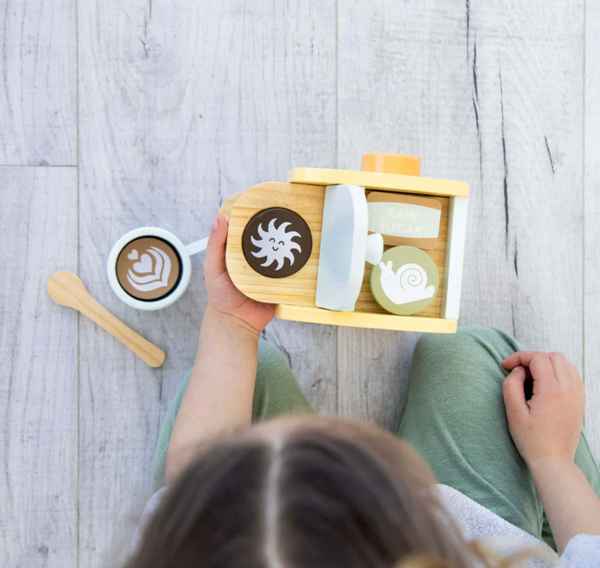 Child playing with the Barista in Training Wooden Coffee Set featuring coffee maker, cup, and spoon on a wooden floor.