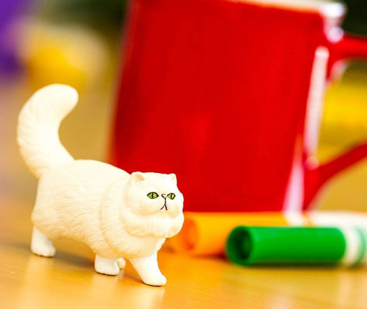 Persian Cat figure with white fluffy coat, peach ears and nose, and green eyes standing near colorful markers and red mug.