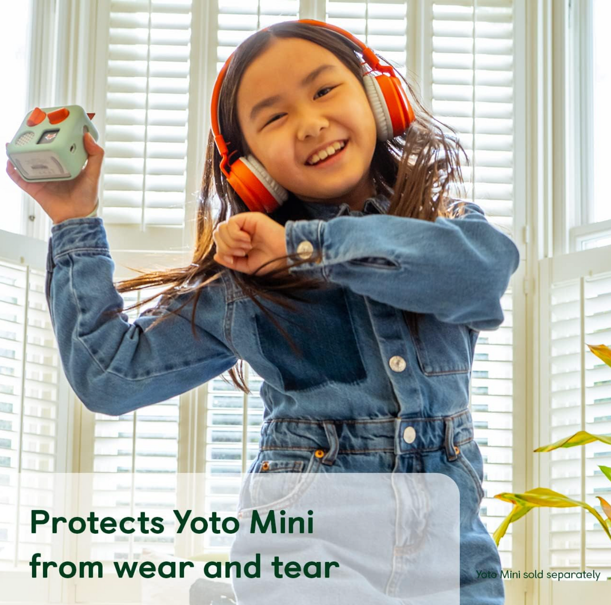 Young girl joyfully holding Yoto Mini protected by Yoto Mini Adventure Jacket | Frog Soup with orange headphones on.