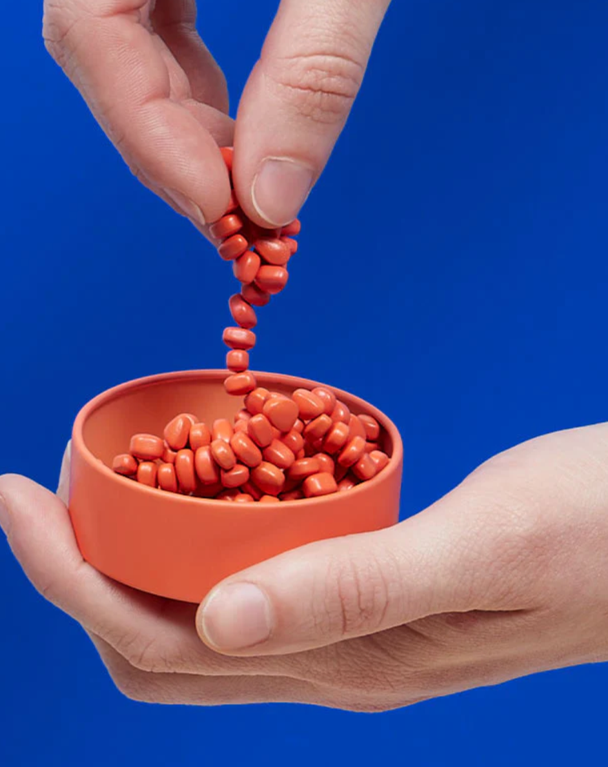 Hands holding and playing with Speks. Matte. Mini Crags. Coral magnetic putty in small coral tin against blue background.
