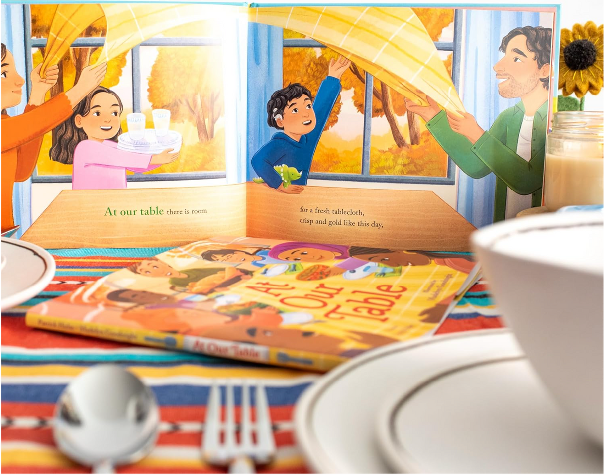 At Our Table book open to colorful illustration of family sharing food at a vibrant Thanksgiving meal.
