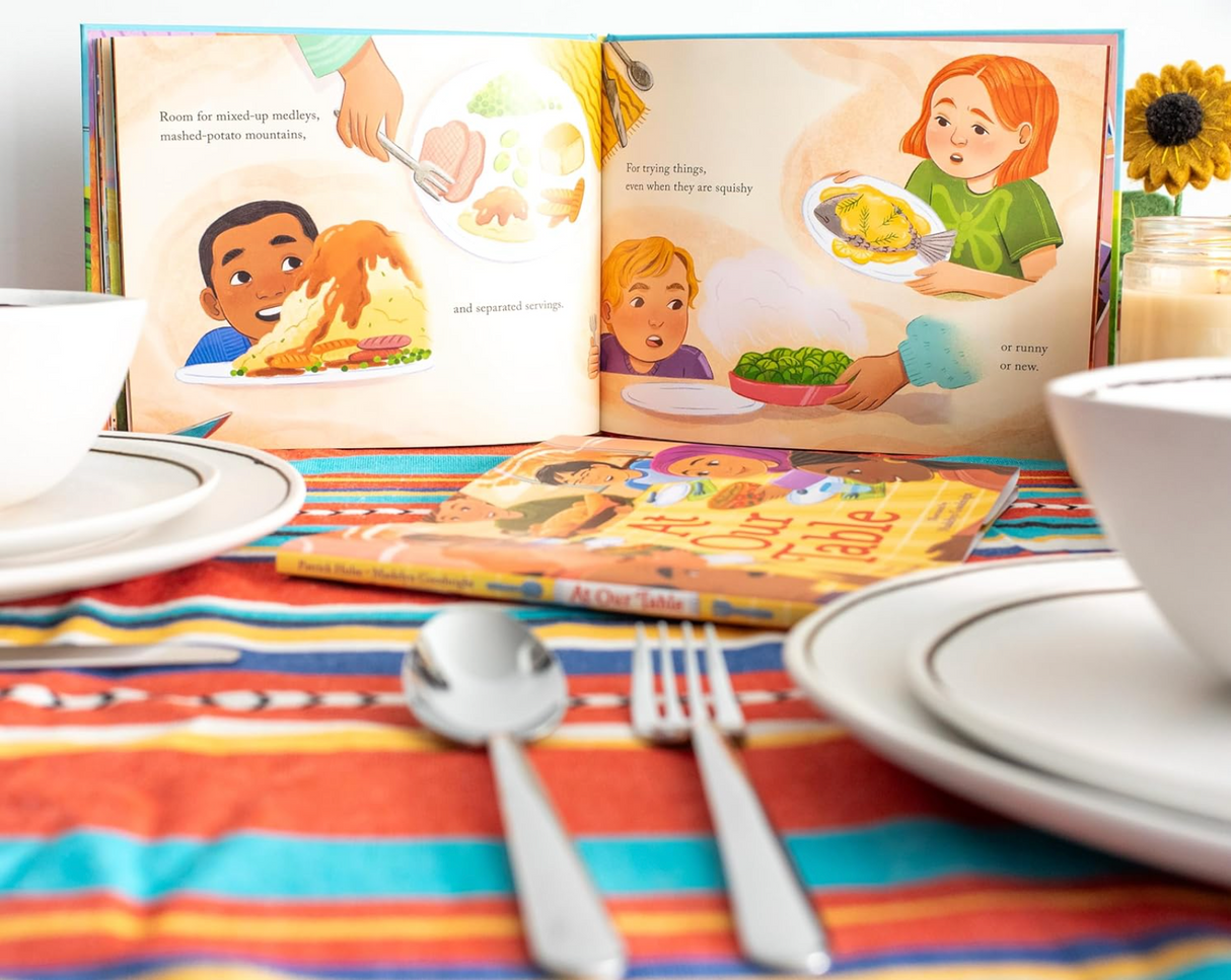 At Our Table book open on colorful tablecloth with plates and utensils, celebrating diverse Thanksgiving traditions.