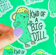 Hand holding "Kind of a Big Dill Sticker" featuring a cool pickle with a hat and shoes on a green background.
