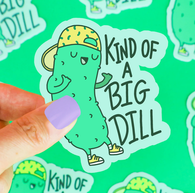 Hand holding "Kind of a Big Dill Sticker" featuring a cool pickle with a hat and shoes on a green background.